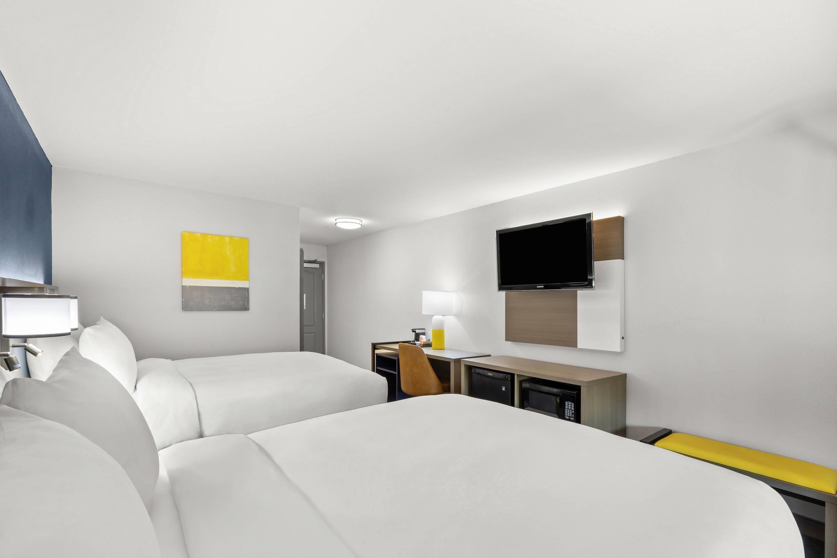 Ett hotellrum med två sängar, ett skrivbord med en stol, en tv som är monterad på väggen och ett litet bord med en lampa.