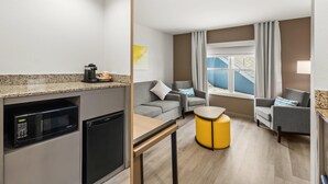 A compact hotel room with a kitchenette, a gray sofa set, a yellow ottoman, and a window with curtains.