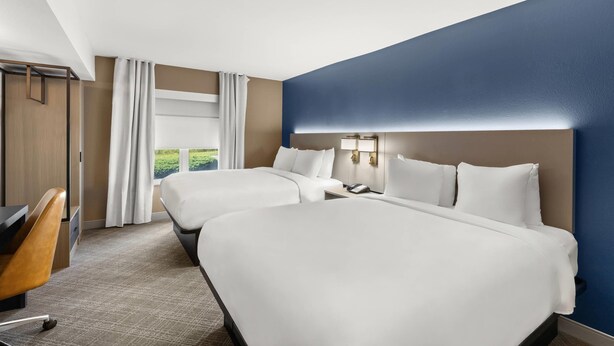 A hotel room with two beds, a desk, and a view of greenery outside.