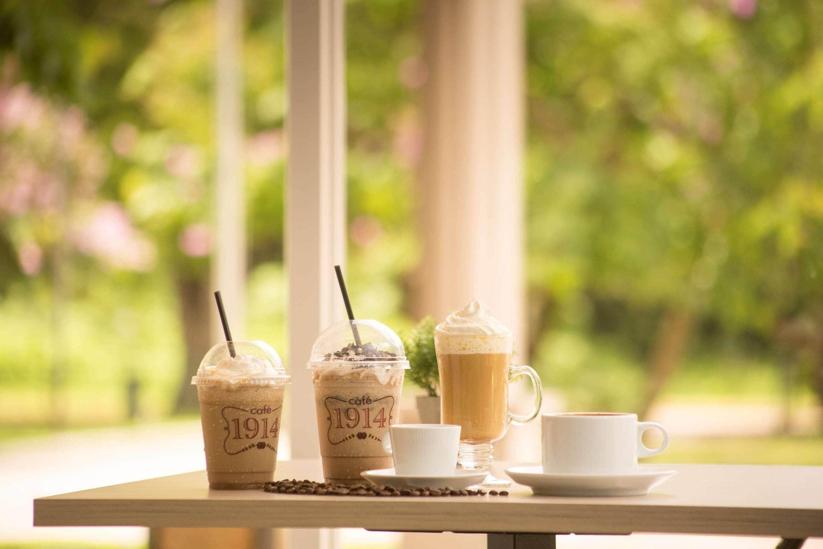 Una mesa con tres cafés helados y una taza blanca, sobre un fondo verde desenfocado.