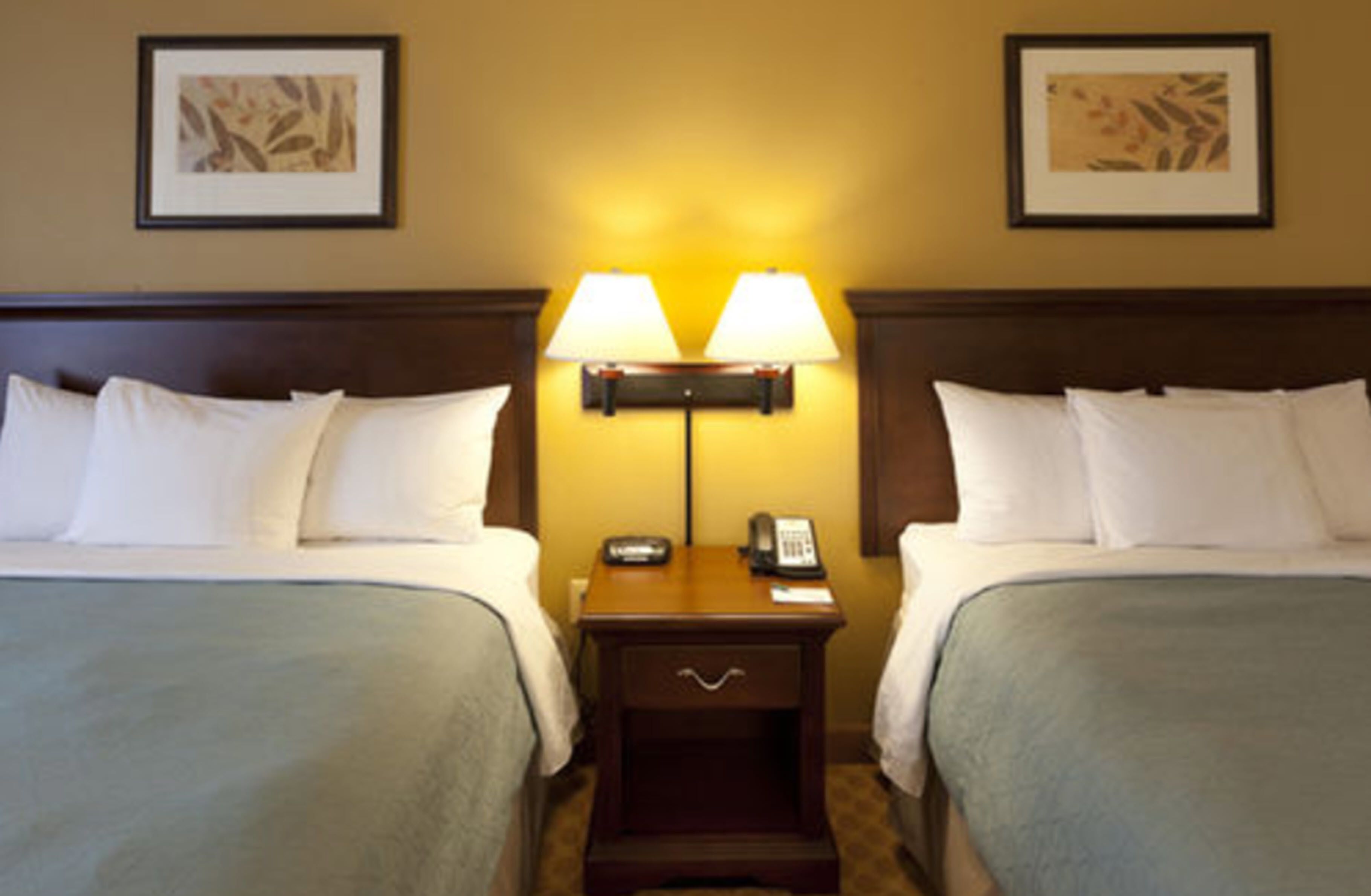 A hotel room with two beds, a nightstand with a lamp, and framed artwork on the walls.