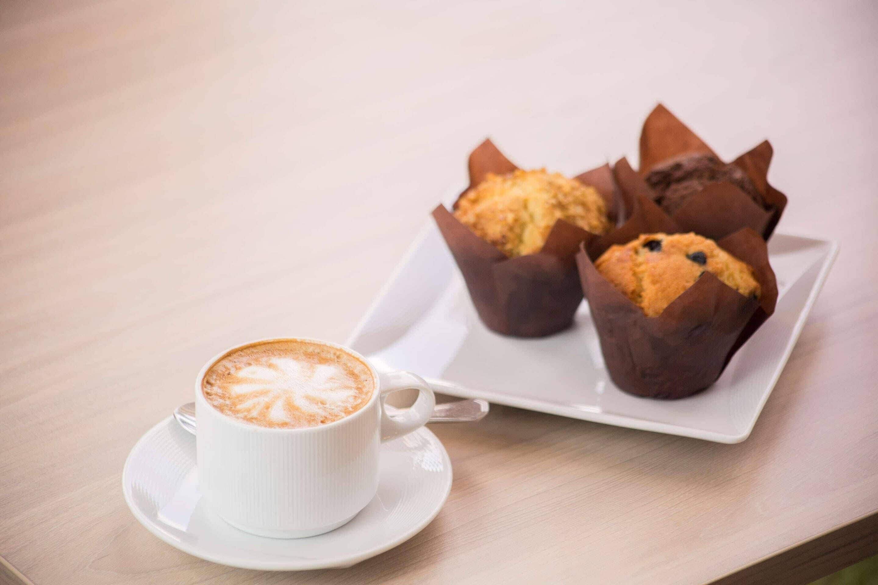 Une tasse de café blanc, décorée d’un motif en latte art, posée sur une soucoupe, accompagnée de deux muffins dans des emballages en papier brun, présentés sur une assiette blanche.