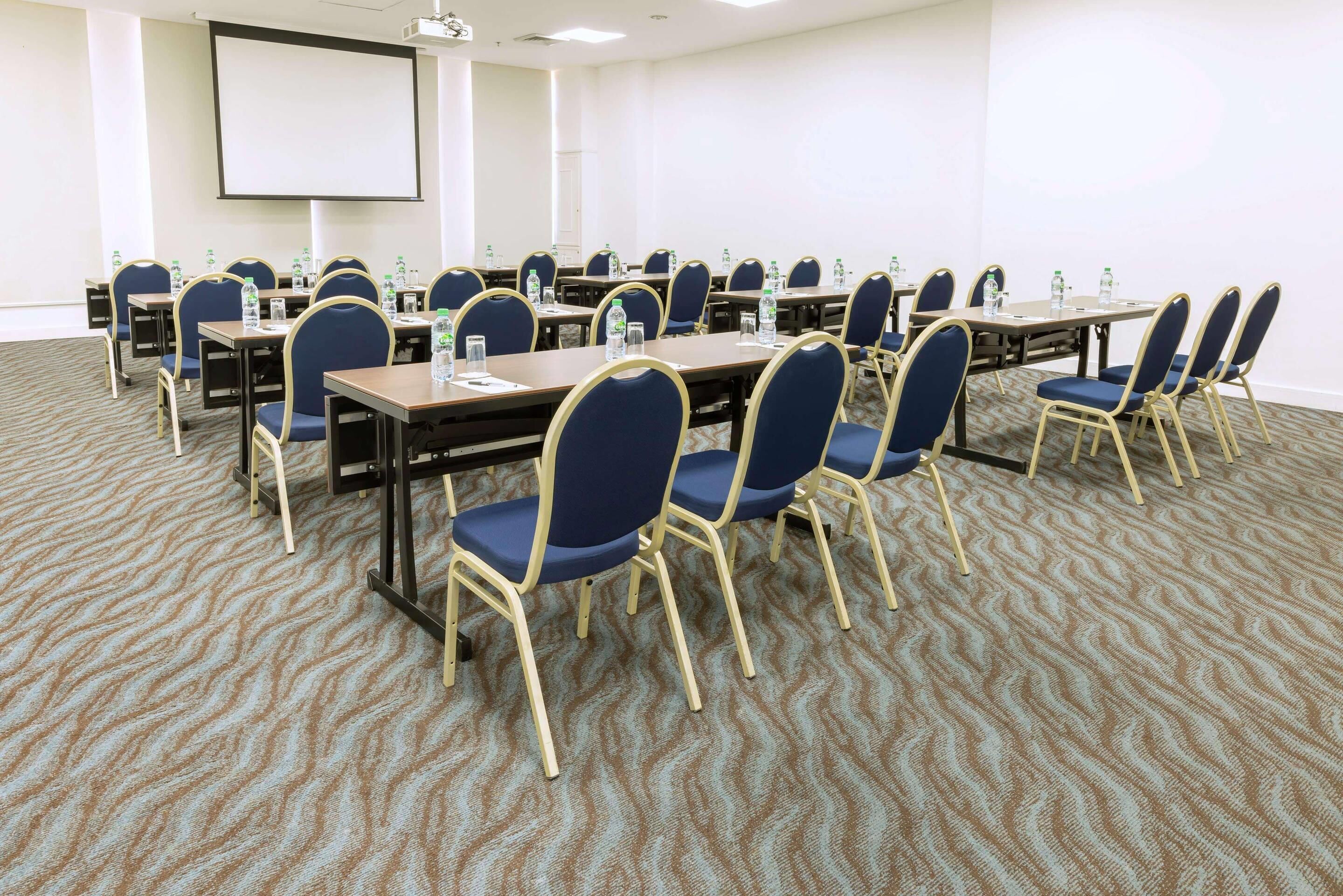 Une salle de conférence dotée de plusieurs tables et de chaises, ainsi que de bouteilles d’eau et d’un écran de projection.