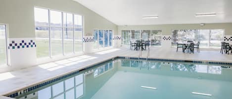 An indoor swimming pool with clear glass walls, a tiled deck, and multiple tables and chairs.
