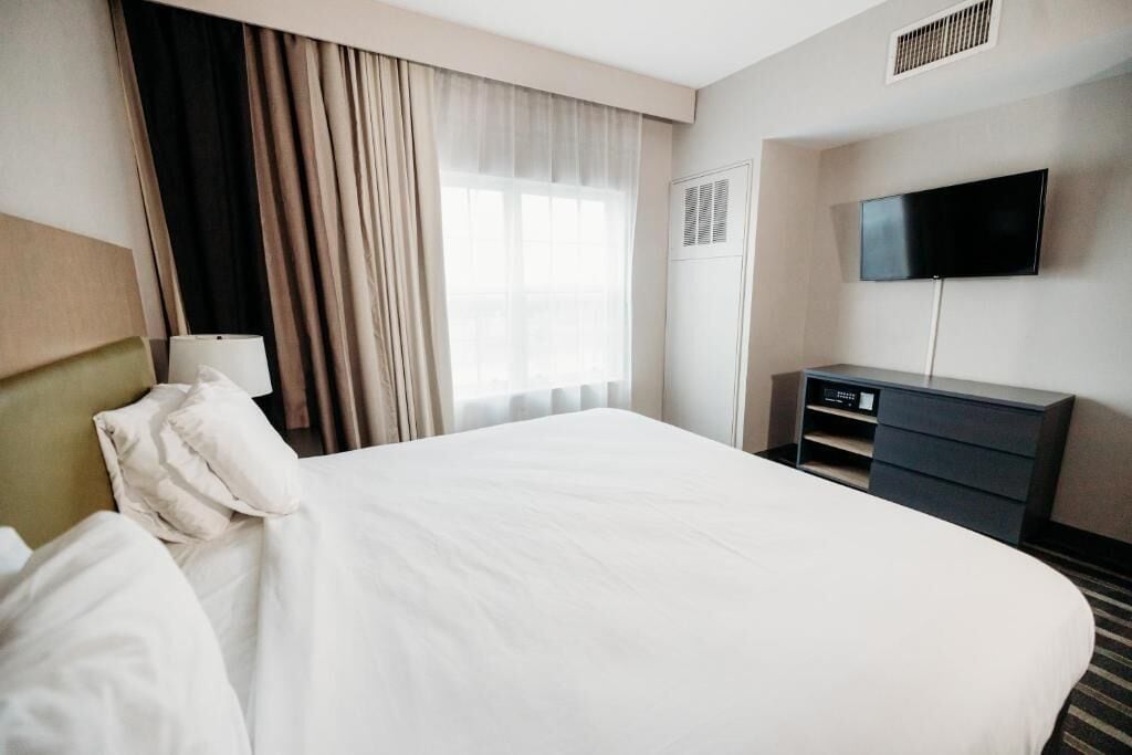 A hotel room with a large bed, a flat-screen TV mounted on the wall, a desk with a lamp, and a window with curtains.