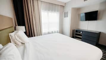 A hotel room with a large bed, a flat-screen TV mounted on the wall, a desk with a lamp, and a window with curtains.