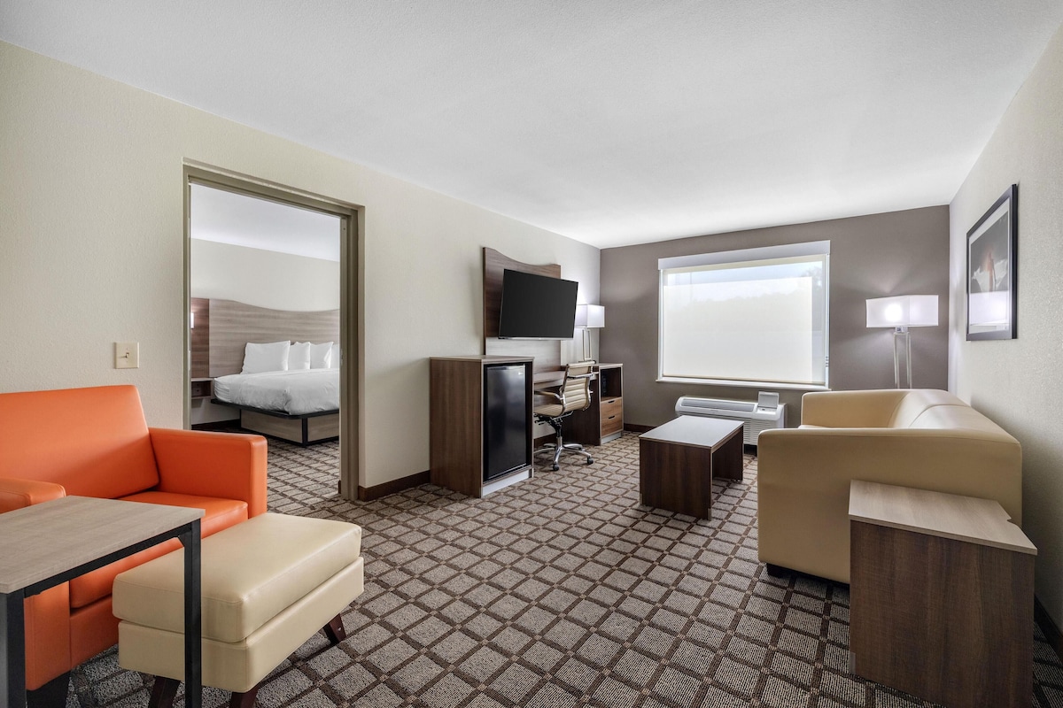 A hotel room with a bed, orange and beige armchairs, a desk, a television, and a window with a view.