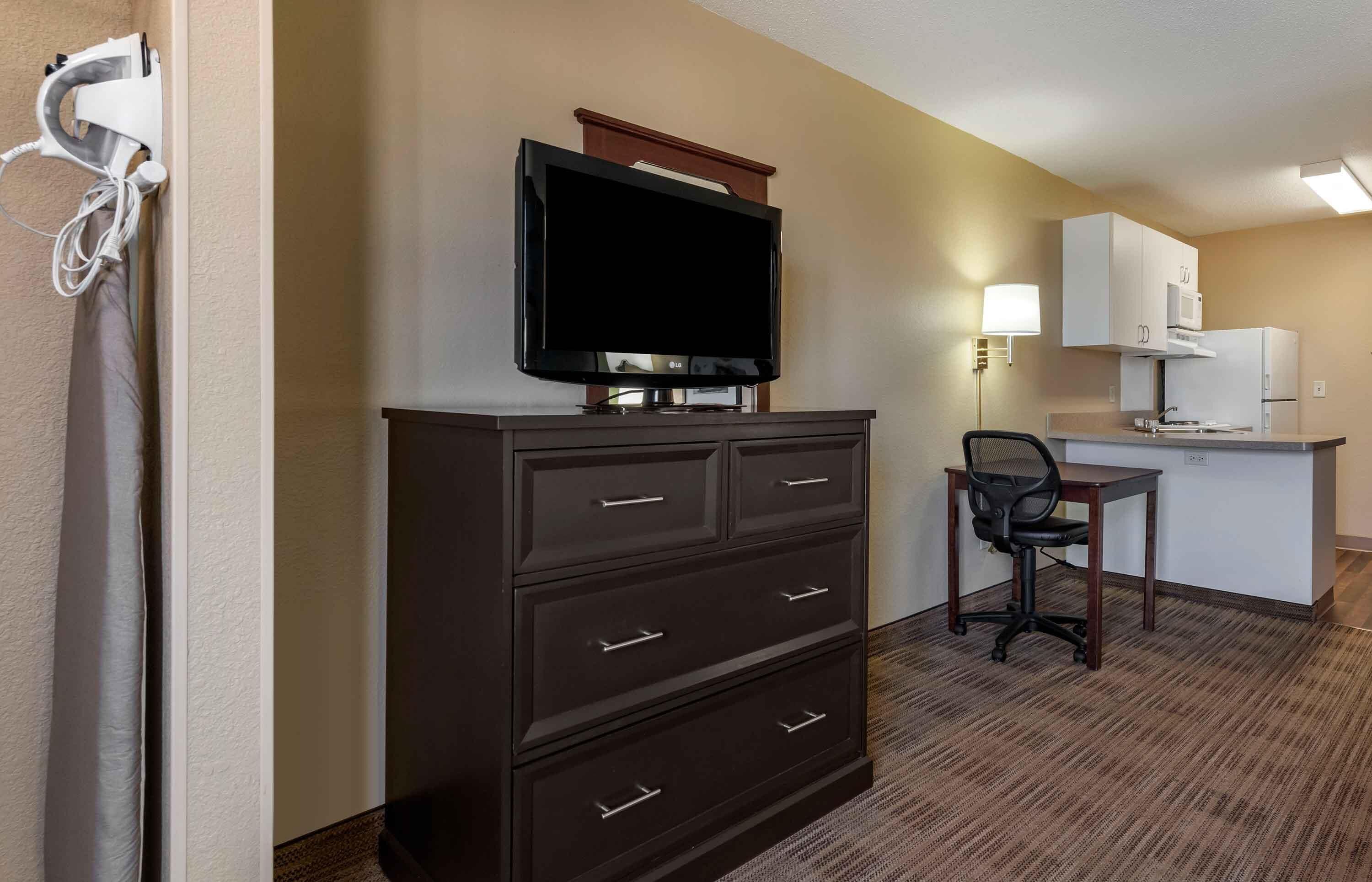 A hotel room with a flat-screen TV on a dresser, a small kitchenette with a microwave, and a chair.