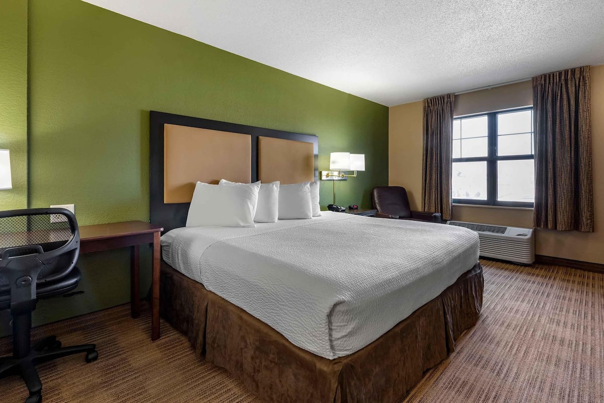 A hotel room with a large bed, a desk with a chair, a lamp, and a window with curtains.
