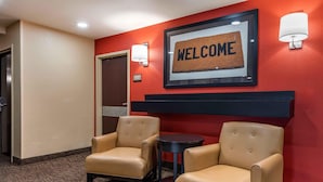 A hotel lobby with two leather armchairs, a small round table, a 'WELCOME' sign, and a 'TRASH CAN' sign on the wall.