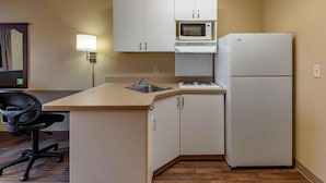 A compact kitchen with white cabinets, a microwave, a refrigerator, a sink, and a chair.