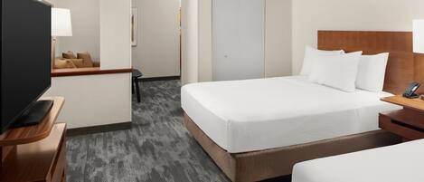 A hotel room with a large bed, a TV on a wooden stand, a desk with a lamp, and a door leading to another room.