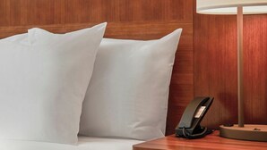 Een hotelkamer met een houten hoofdeinde, wit beddengoed, een bedlampje en een telefoon op het nachtkastje.