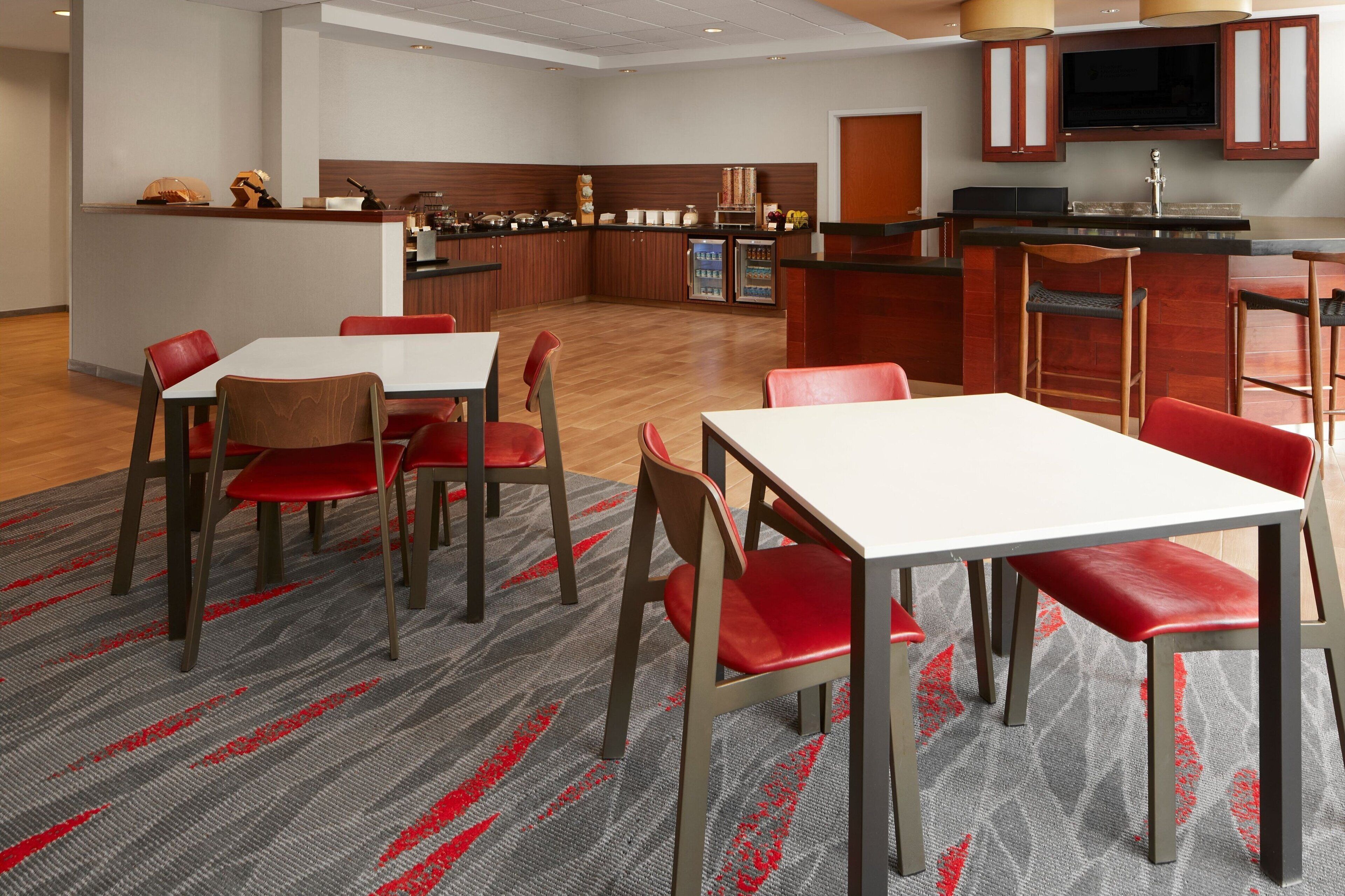 A modern hotel dining area with a central island, red upholstered chairs, and a grey patterned carpet.