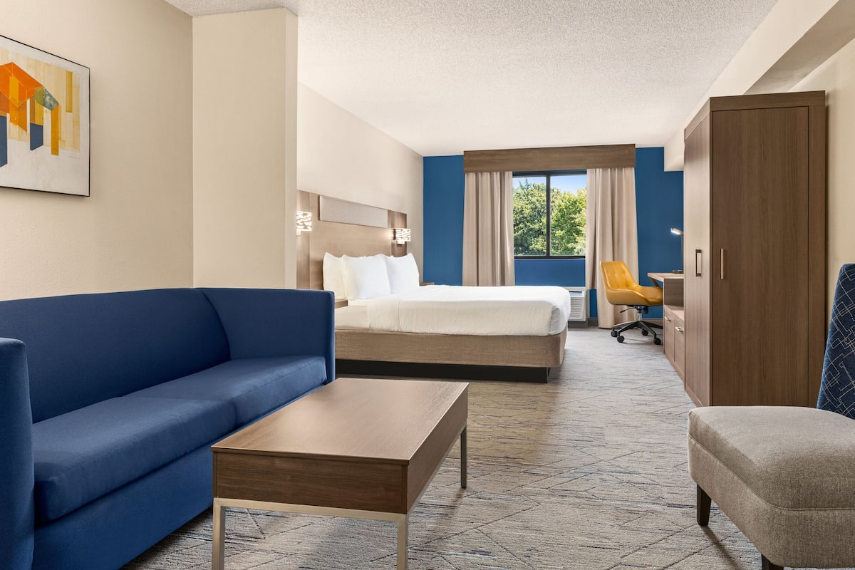 Habitación de hotel con un sofá azul, una mesa de centro de madera, una cama con ropa de cama blanca, un armario de madera y un escritorio con una silla.
