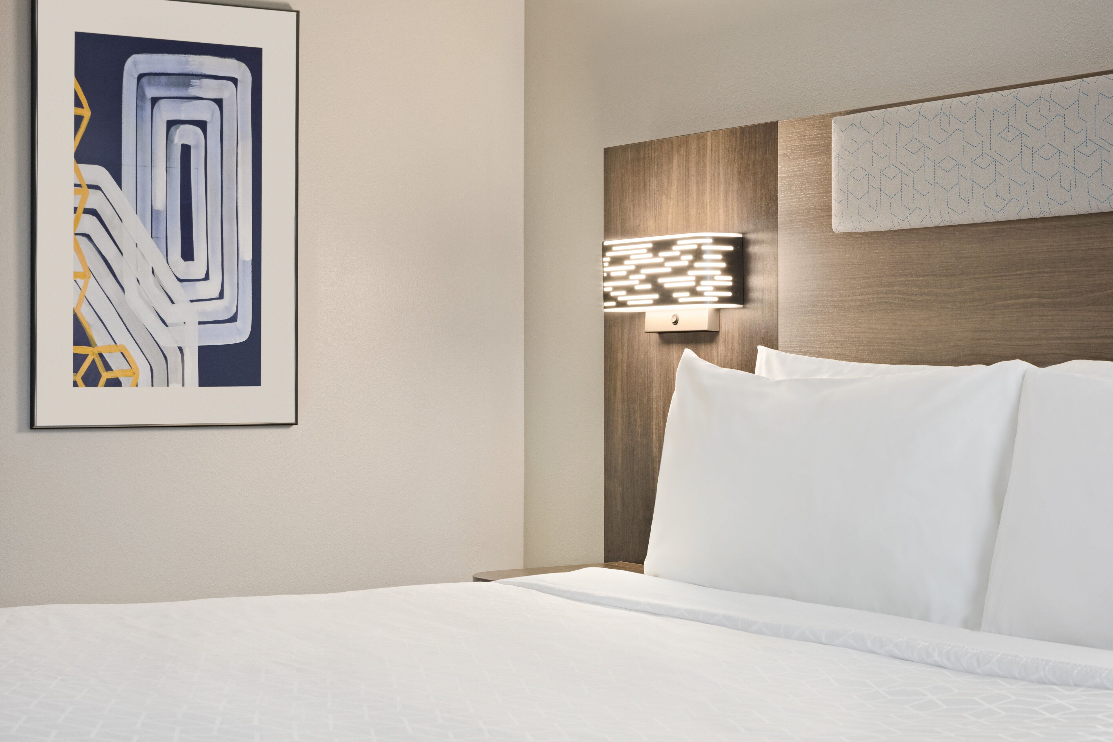 Una habitación de hotel con una cama bien hecha, una obra de arte moderna en la pared y una lámpara fijada a la pared.