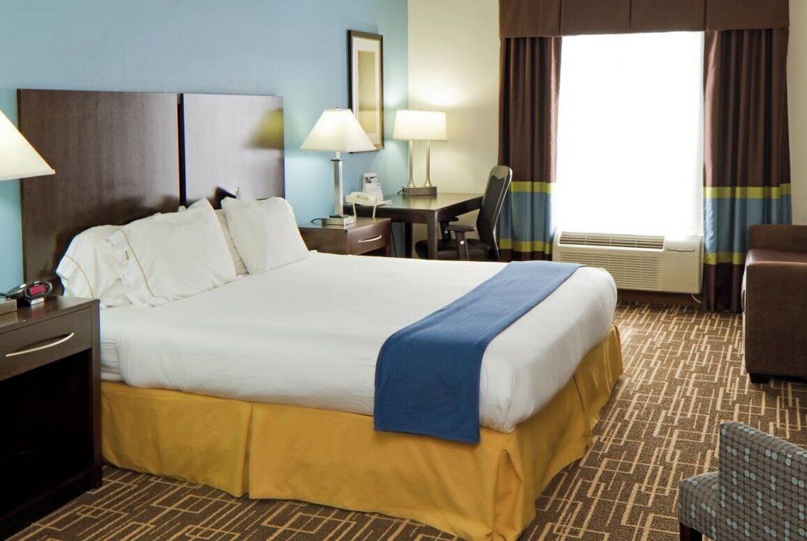 Habitación de hotel con una cama grande, un escritorio, una silla, una lámpara y un ventanal con cortinas.