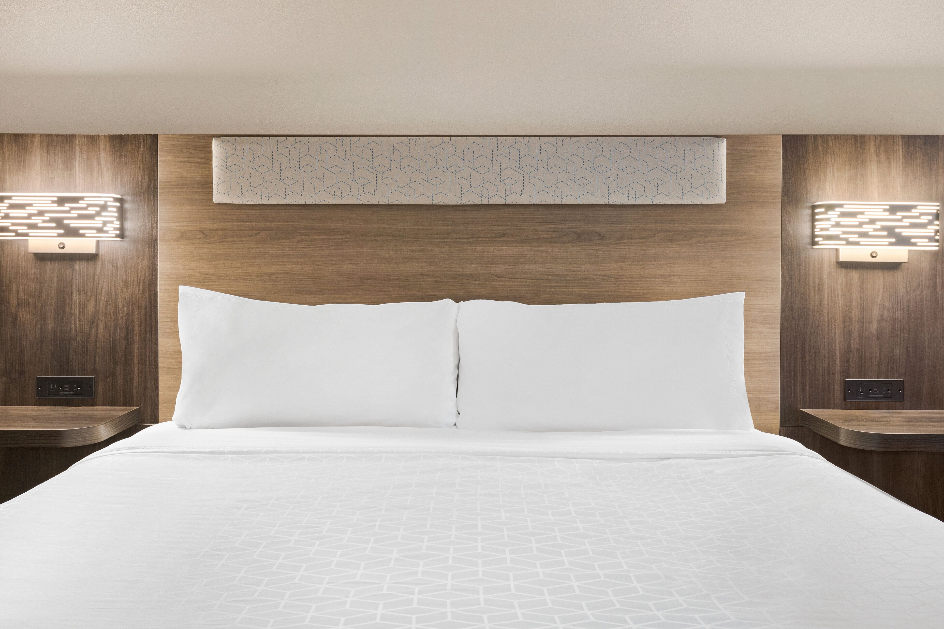 Una habitación de hotel con una cama grande, dos mesitas de noche, una lámpara de pared y un enchufe.