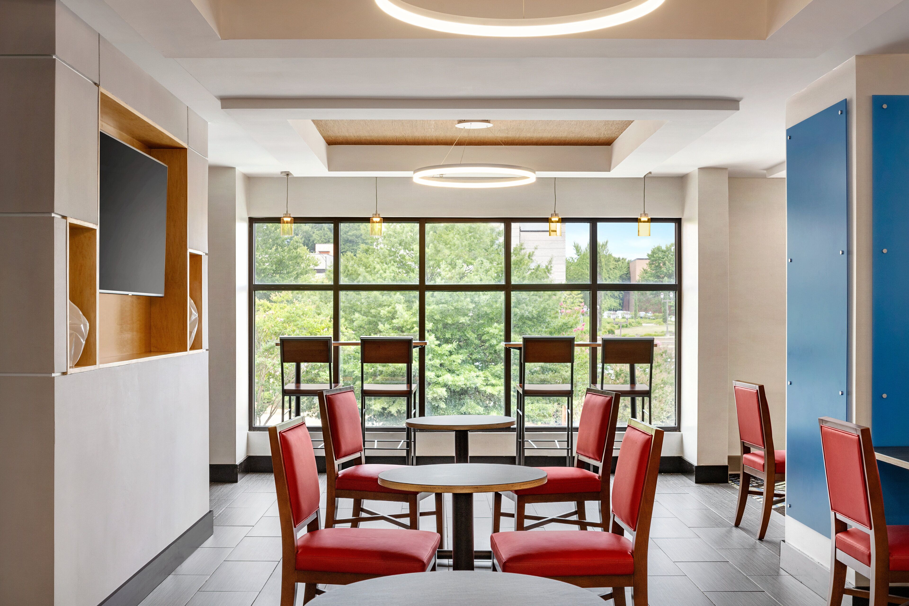 A modern interior with a round table and red chairs, large windows with a view of greenery, and a blue door.