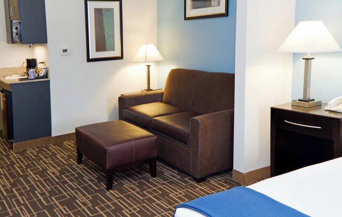 A hotel room with a bed, a sofa, an ottoman, a desk with a lamp, and a small kitchenette area.