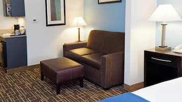 A hotel room with a bed, a sofa, an ottoman, a desk with a lamp, and a small kitchenette area.