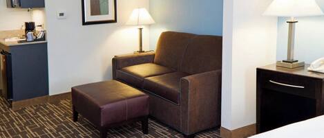 A hotel room with a bed, a sofa, an ottoman, a desk with a lamp, and a small kitchenette area.