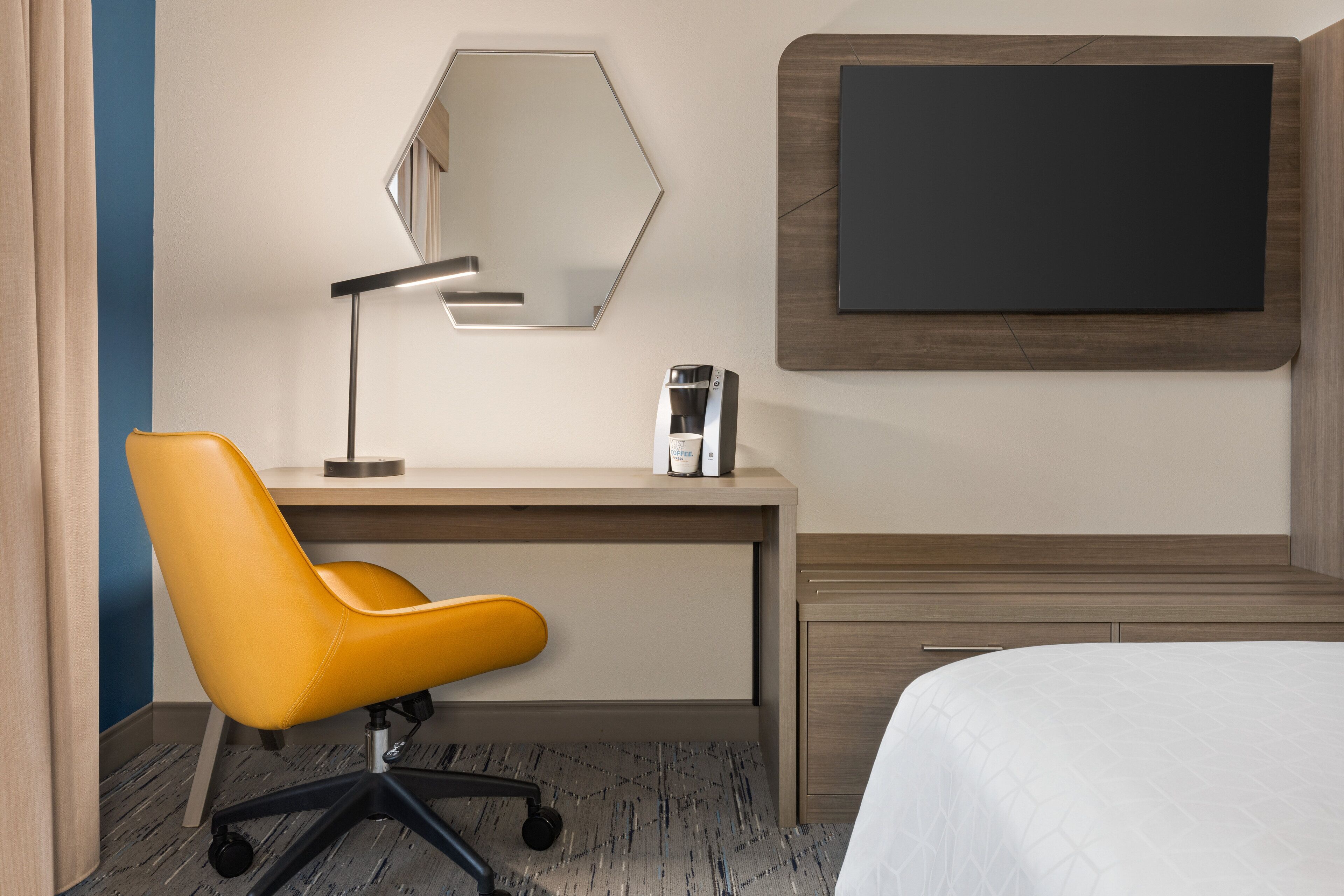 A hotel room with a wooden desk, a yellow office chair, a bedside table, a bed with a white bedspread, a wall-mounted TV, and a mirror.