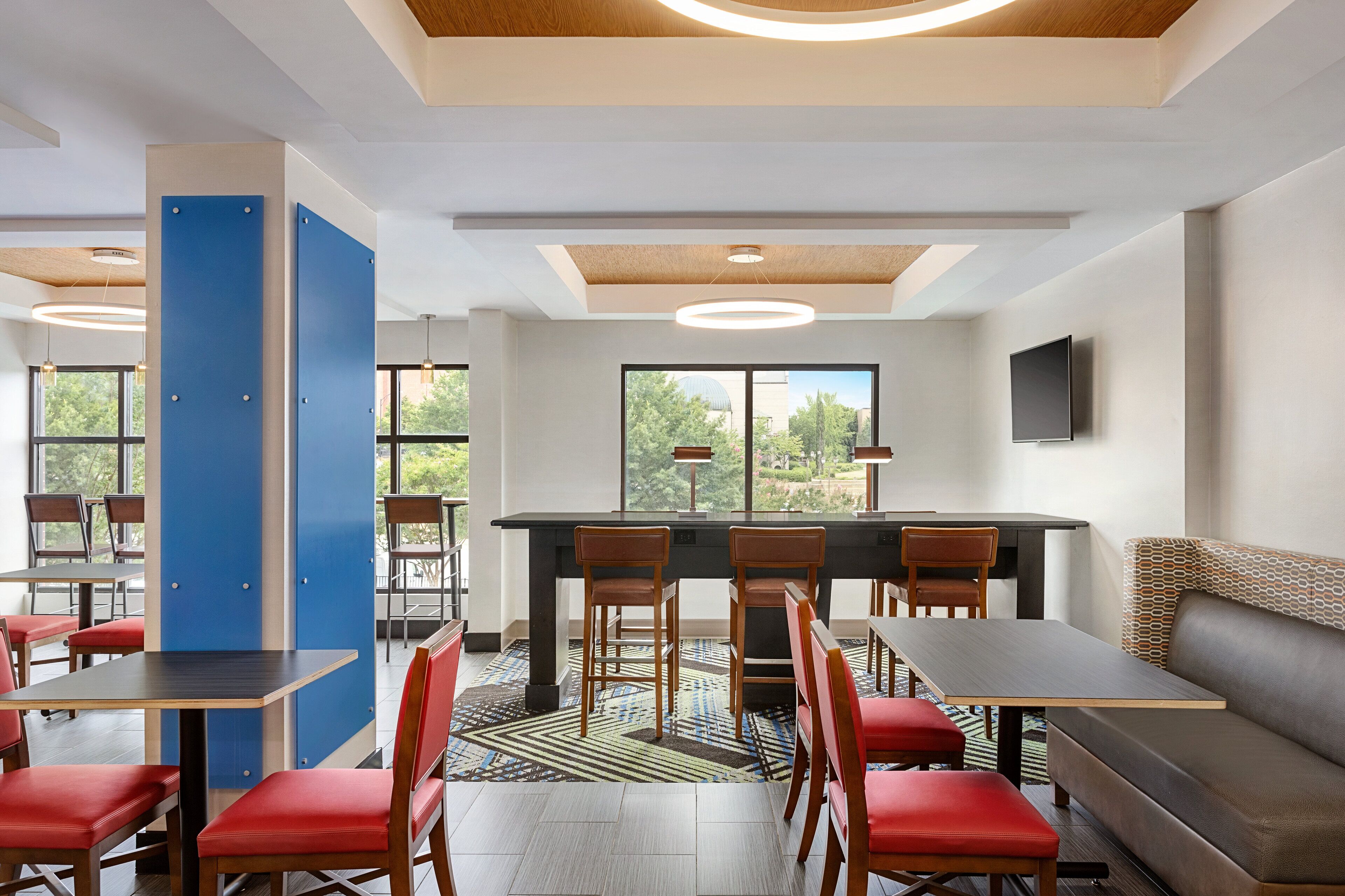 Un hall d’hôtel moderne comprenant un espace repas, plusieurs tables et chaises, un canapé et de grandes fenêtres donnant sur un paysage verdoyant.