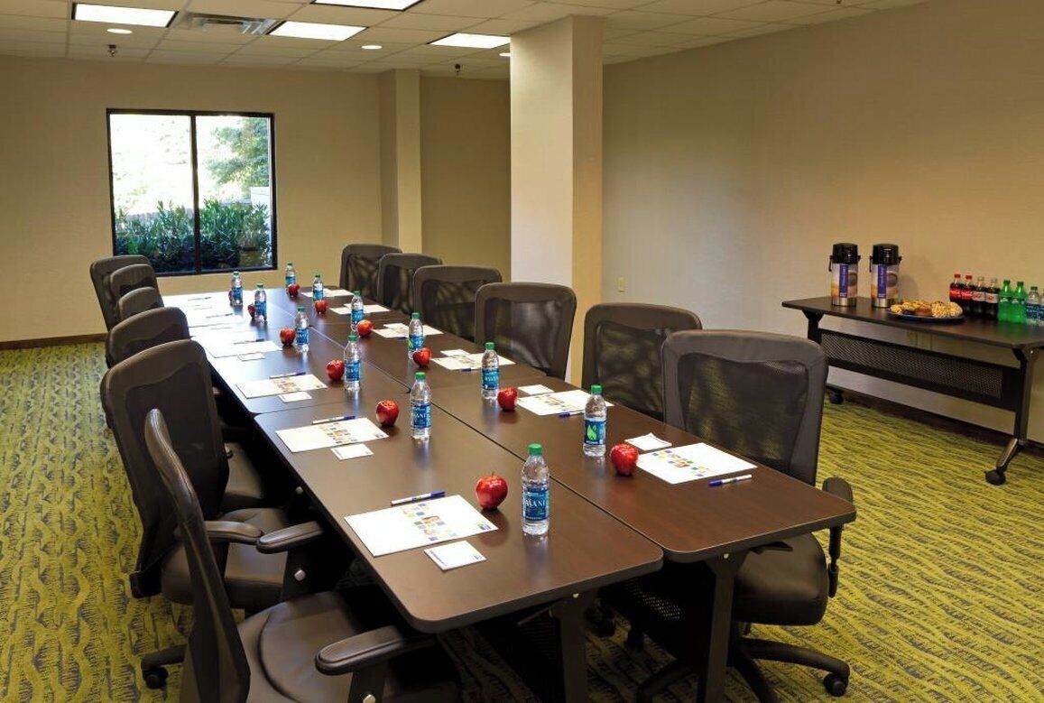Una sala de conferencias con una larga mesa, sillas, botellas de agua, manzanas y una mesa con agua embotellada y refrigerios.
