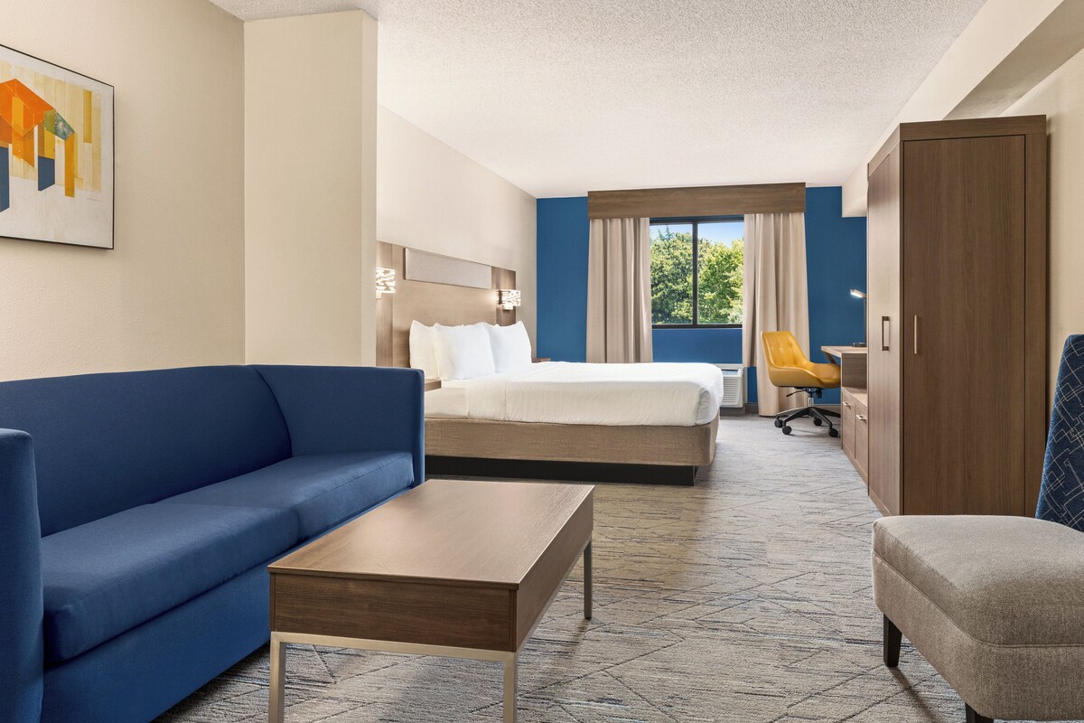 Habitación de hotel con un sofá azul, una mesa de centro de madera, una cama con ropa de cama blanca, un armario de madera y un escritorio con una silla.