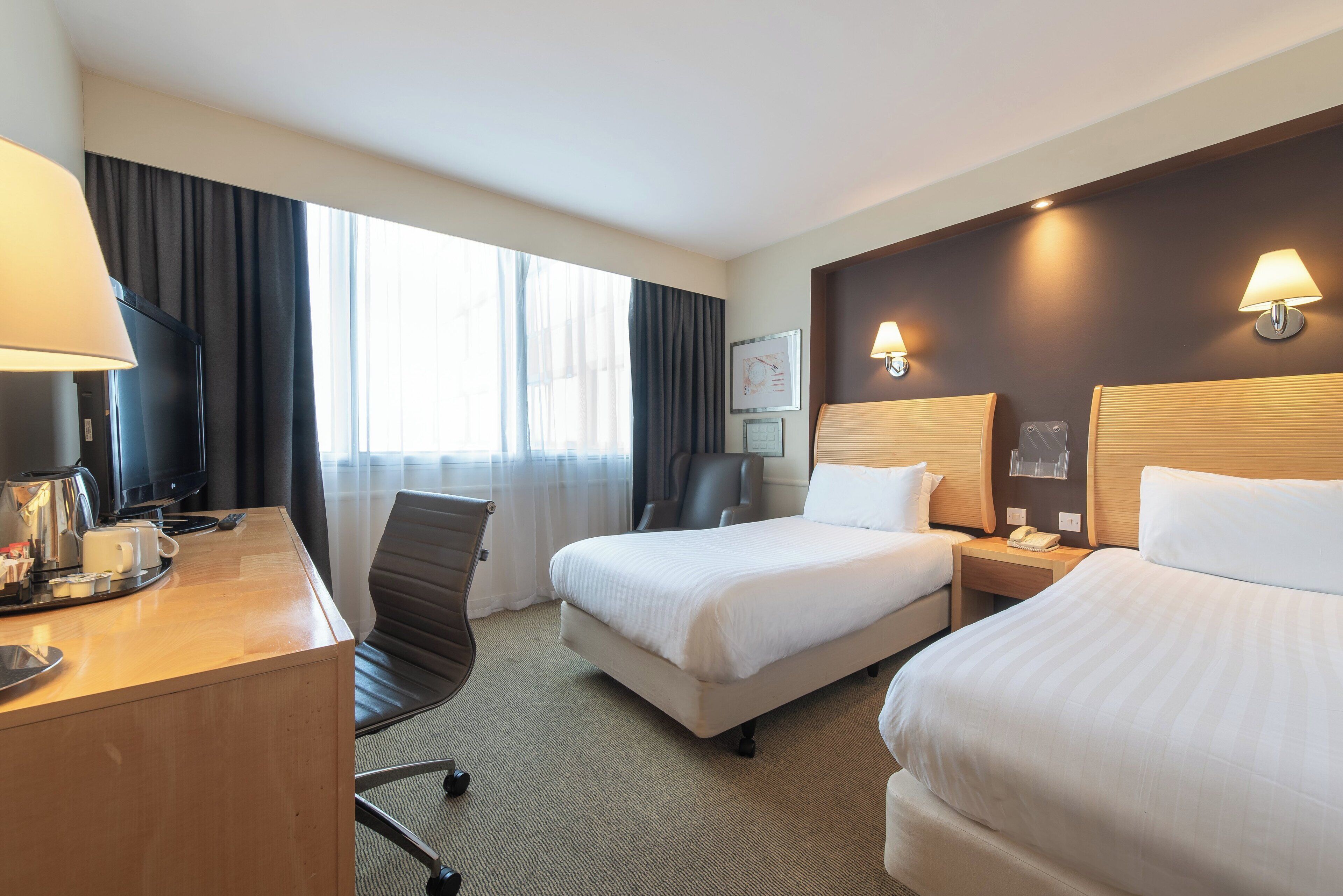 A hotel room with two beds, a desk with a coffee maker, a TV, and a large window with curtains.