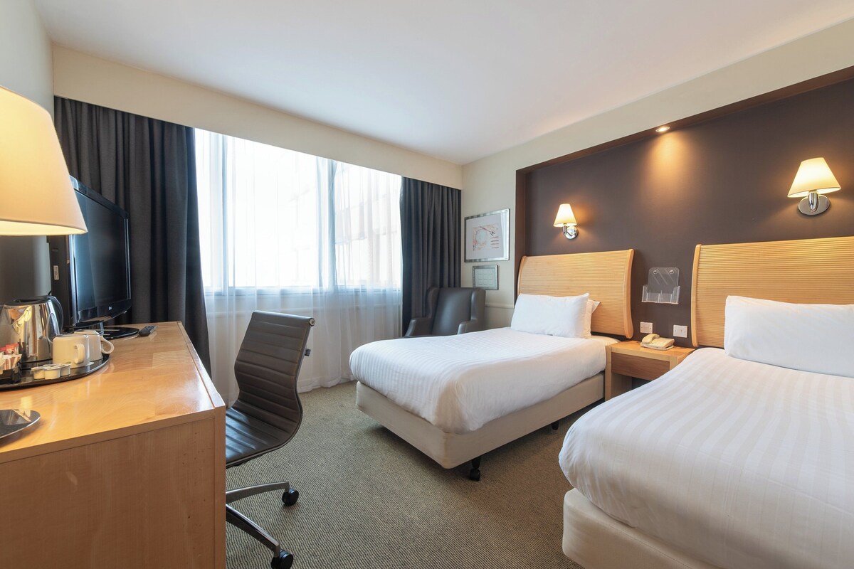 Habitación de hotel con dos camas, un escritorio con cafetera, televisión y un amplio ventanal con cortinas.