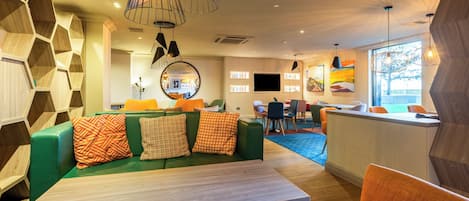 A modern lounge area with a green sofa, orange chairs, a wooden table, and a large window providing natural light.