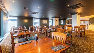 A spacious restaurant with multiple wooden tables and chairs, large windows, and a patterned carpet.