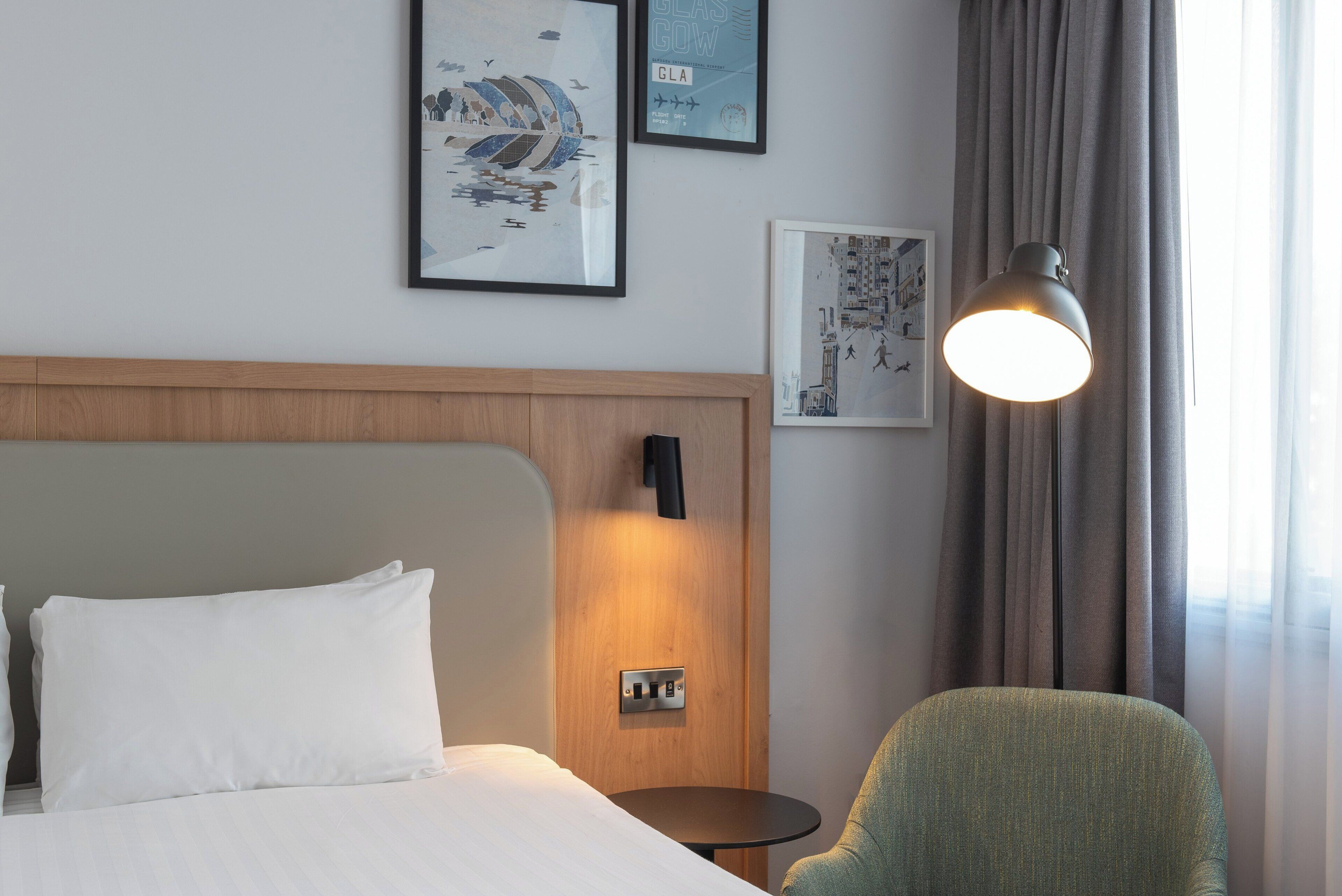 A hotel room with a bed, a bedside table, a lamp, and a chair. There are two framed pictures on the wall.