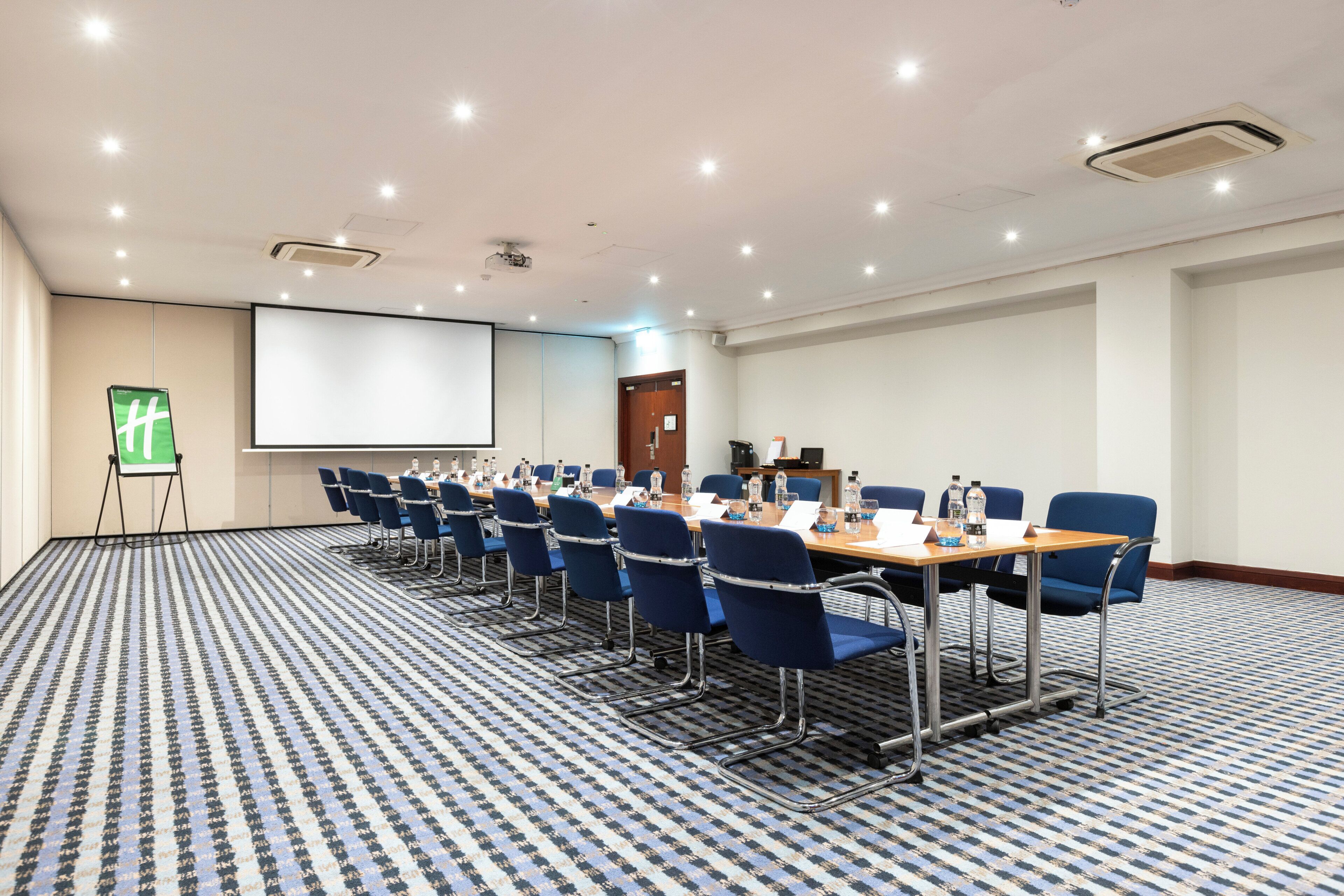Un salón de conferencias con una gran mesa, sillas azules, una pantalla de proyector y piso alfombrado.