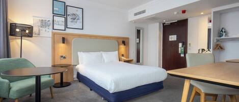 A hotel room with a bed, a desk, a chair, a lamp, and framed pictures on the wall.