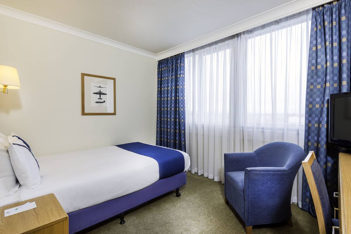 Une chambre d’hôtel avec un lit, une chaise, un bureau, une lampe, une télévision et une fenêtre avec des rideaux.