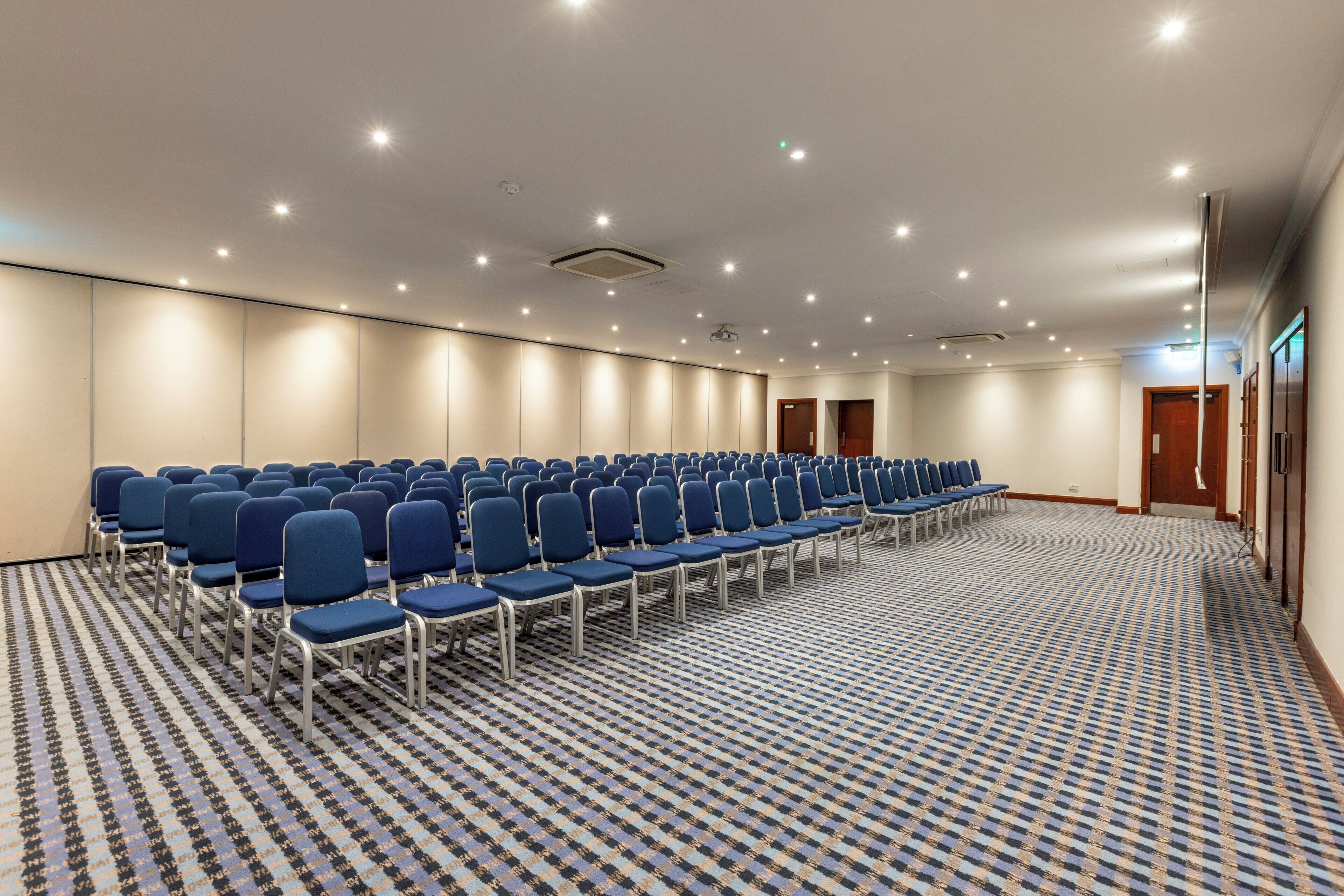 A conference room with rows of blue chairs, a patterned carpet, and recessed ceiling lights.