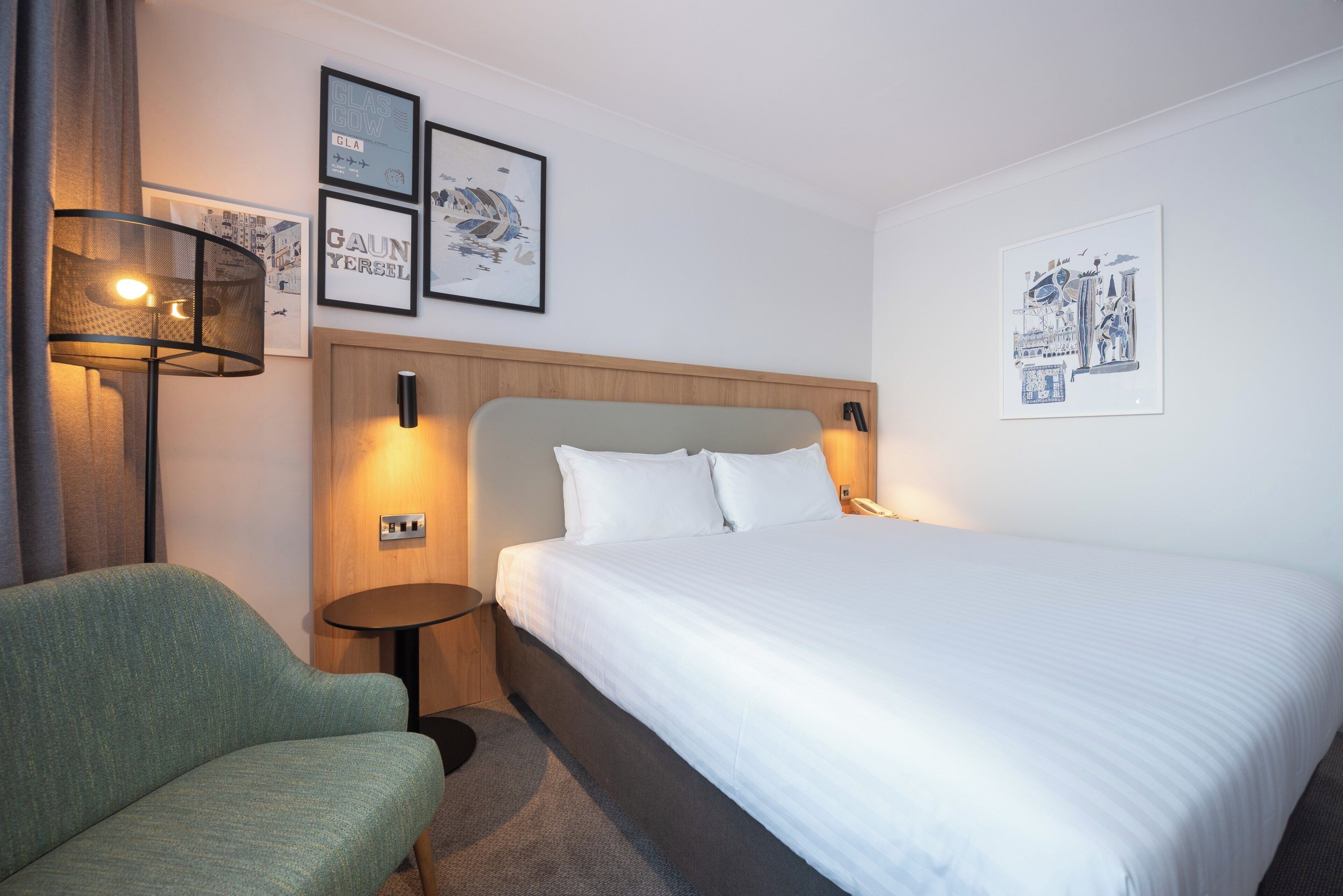 Une chambre d’hôtel avec un lit, un fauteuil vert, une table de chevet et des œuvres d’art murales.