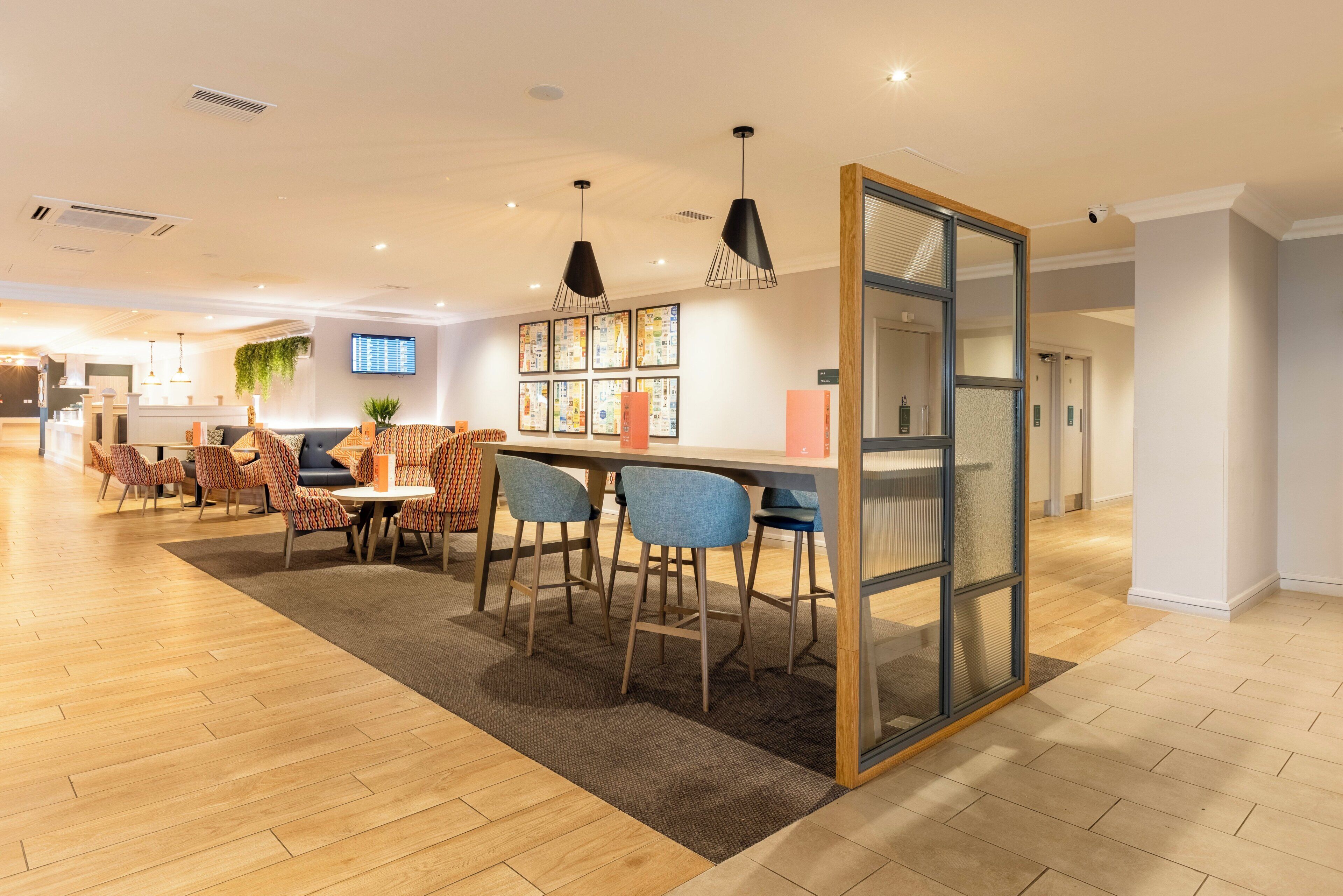 A modern hotel lobby with a wooden floor, a reception desk, a seating area with patterned chairs, and a bar with stools.