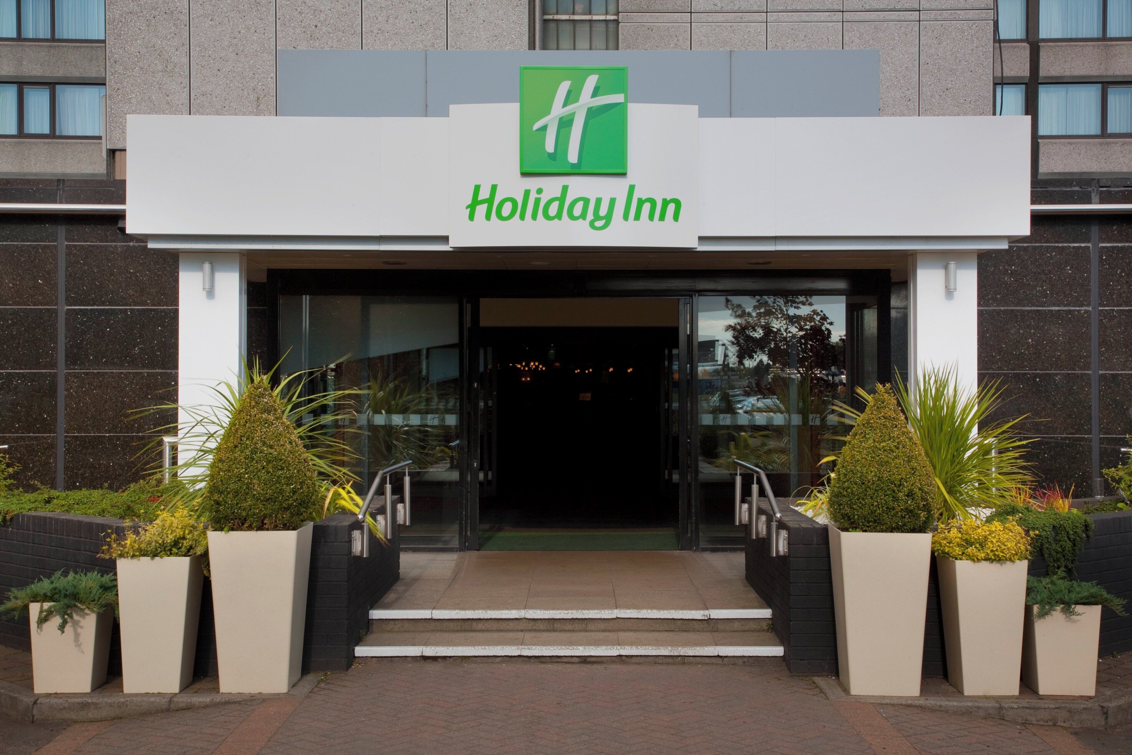La entrada a un Holiday Inn con logotipo verde, una serie de escalones que conducen a la entrada y plantas en macetas a ambos lados.
