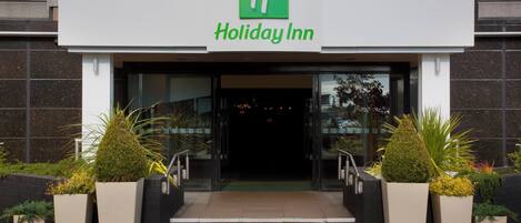 La entrada a un Holiday Inn con logotipo verde, una serie de escalones que conducen a la entrada y plantas en macetas a ambos lados.