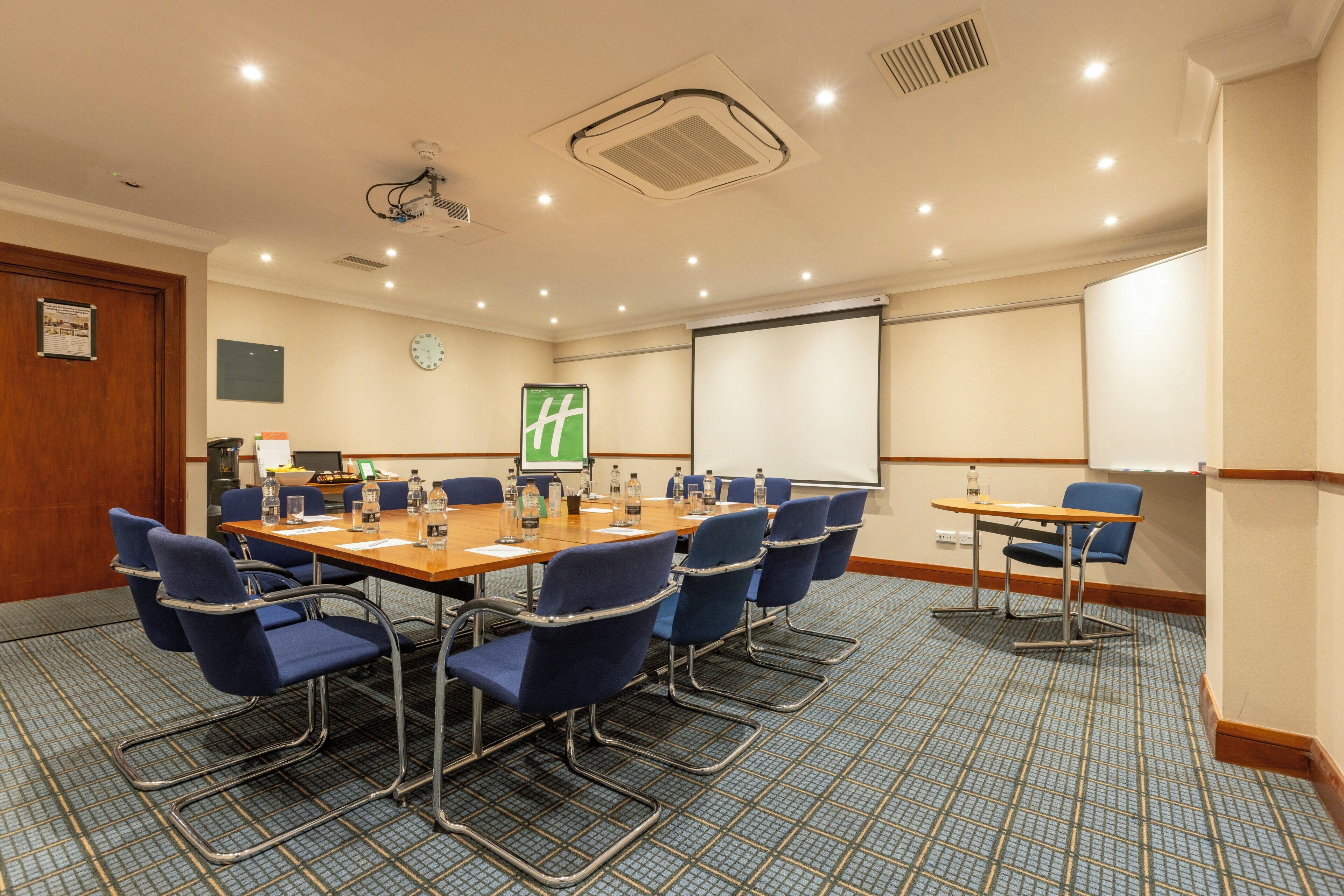 A conference room with a large table, blue chairs, a projector screen, and a clock on the wall.