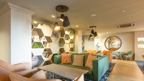 A modern lobby with a green sofa, orange chairs, a wooden coffee table, and geometric wall shelving.