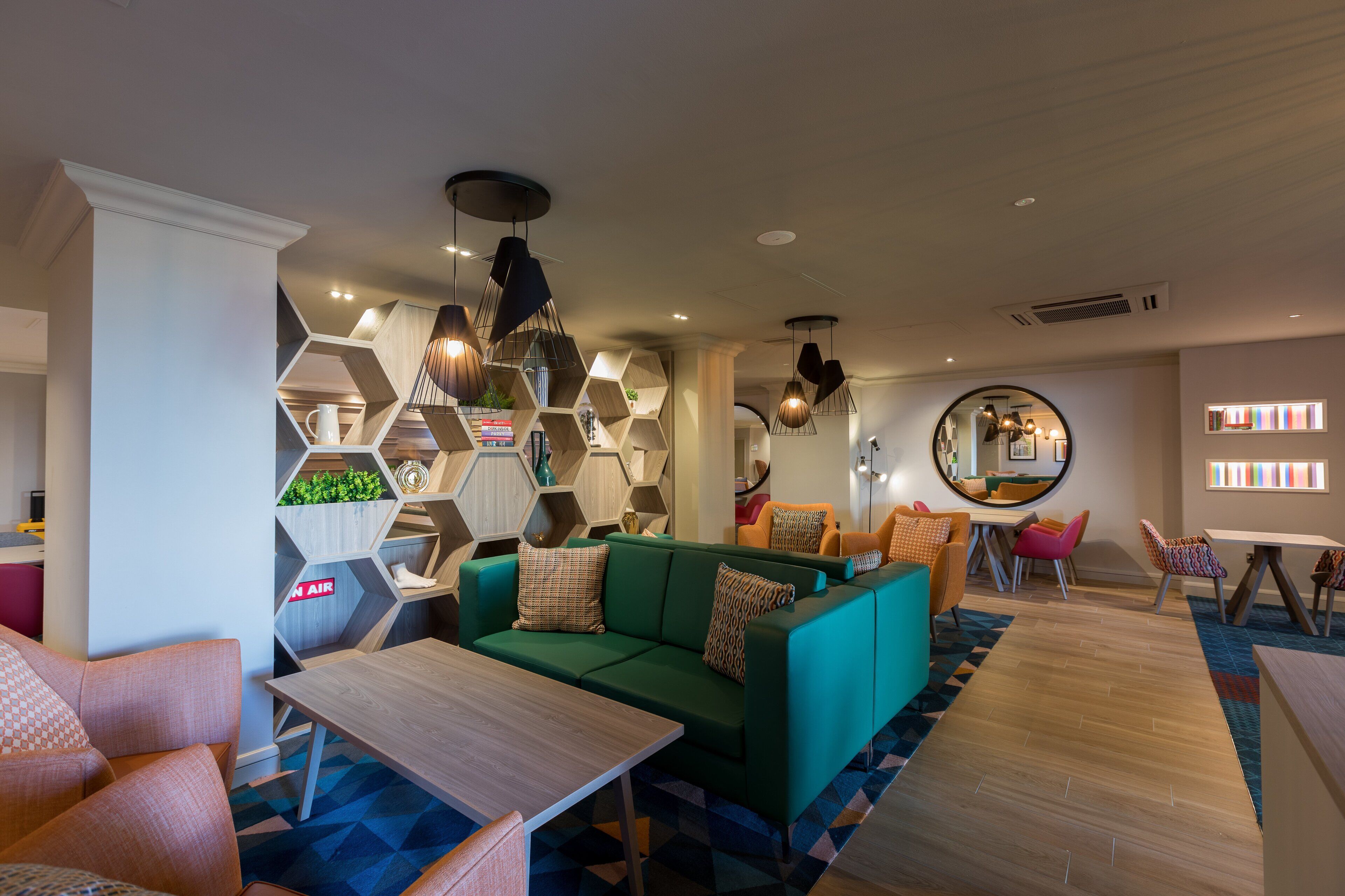 Un espace de détente moderne avec un canapé vert, une table basse en bois et des chaises colorées. On y trouve des étagères hexagonales ornées de plantes et de livres, ainsi qu’un miroir rond accroché au mur.