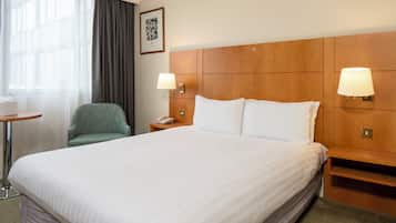 Habitación de hotel con una cama grande, dos lámparas de noche, una silla, una mesita y un cuadro en la pared.