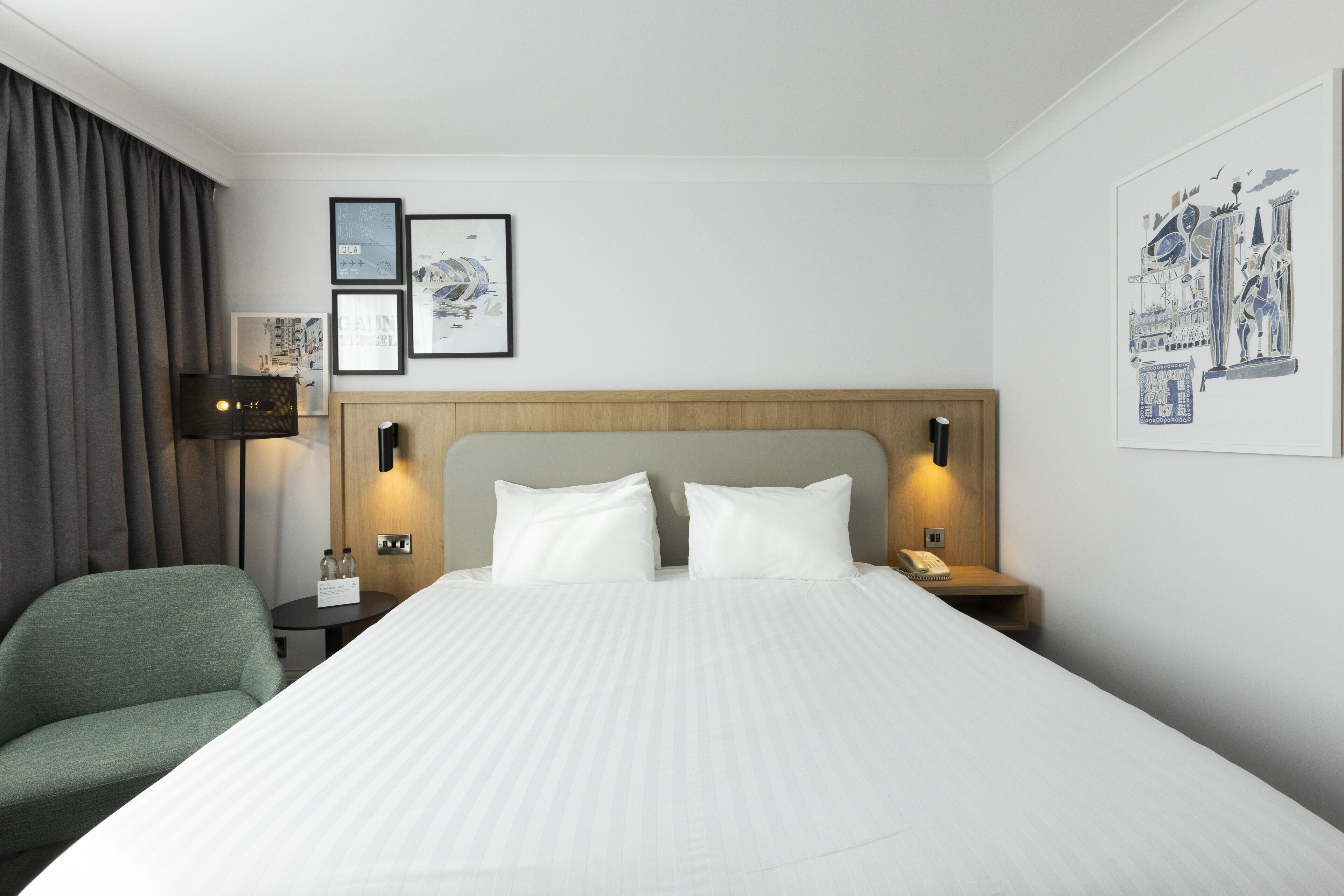 A hotel room with a large bed, a green armchair, a bedside table with a lamp, and framed artwork on the wall.