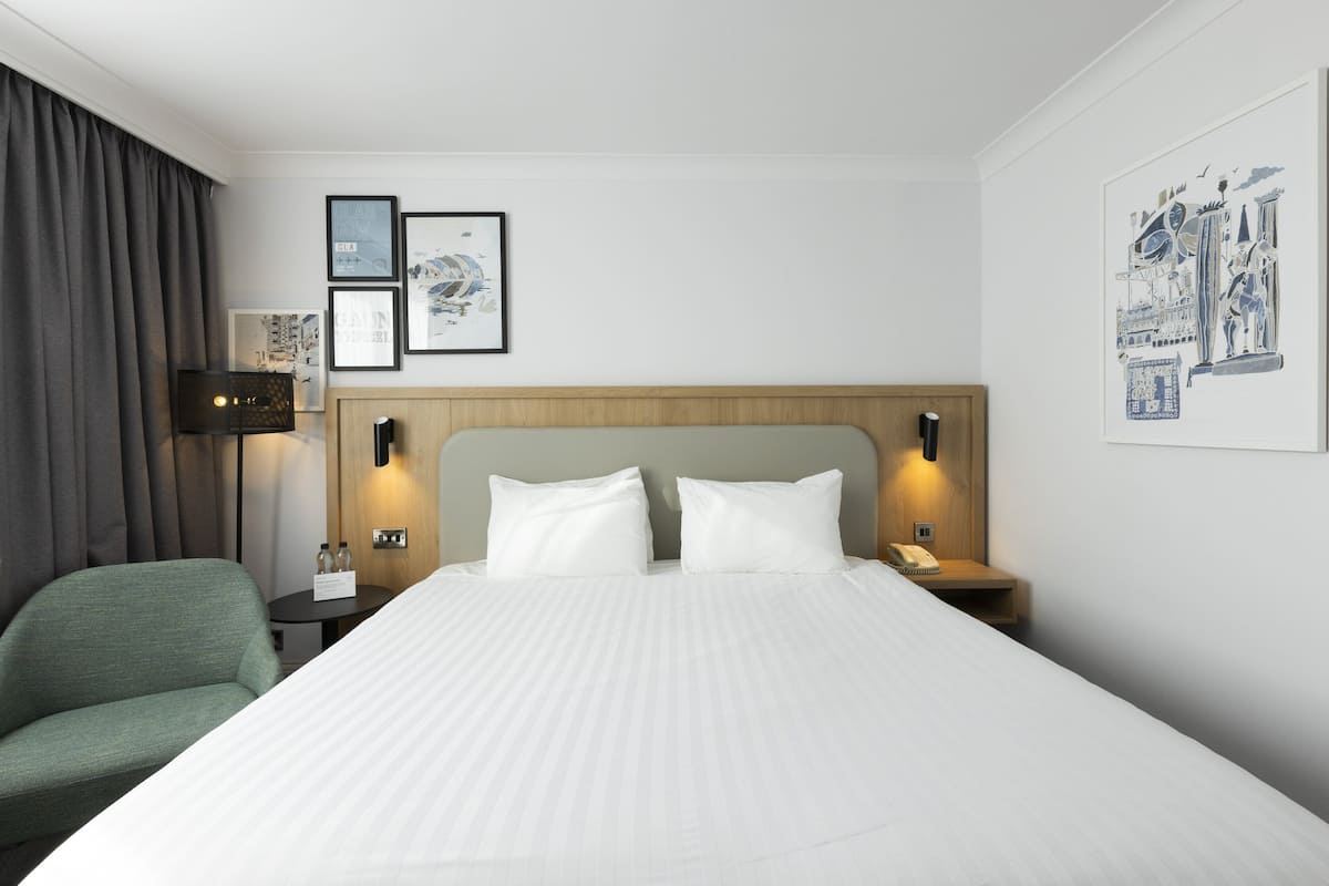 A hotel room with a large bed, a green armchair, a bedside table with a lamp, and framed artwork on the wall.