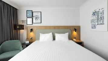 Una habitación de hotel con una cama grande, un sillón verde, una mesita de noche con una lámpara y cuadros en la pared.